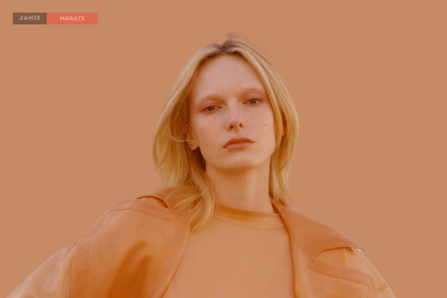 A portrait of a blonde model wearing a monochrome outfit against a warm-toned background.