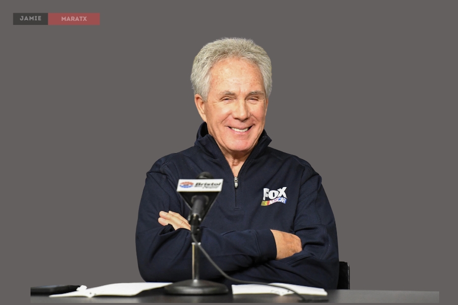 Darrell Waltrip at a press conference, wearing a Fox NASCAR jacket and smiling.