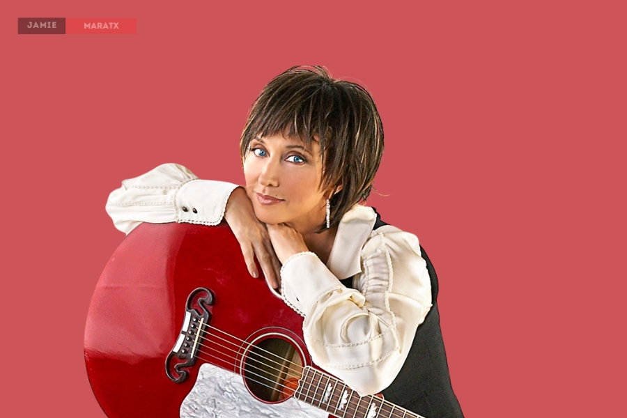 Pam Tillis posing with a red acoustic guitar against a solid red background.