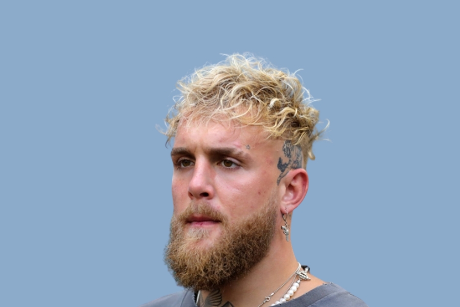 Jake Paul with blond curly hair and beard, wearing jewelry against a blue background.