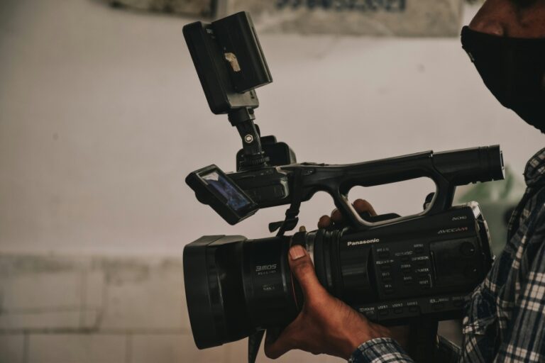 Close-up of a war correspondent holding a professional video camera, wearing a mask and flannel shirt, preparing to film in a conflict zone.
